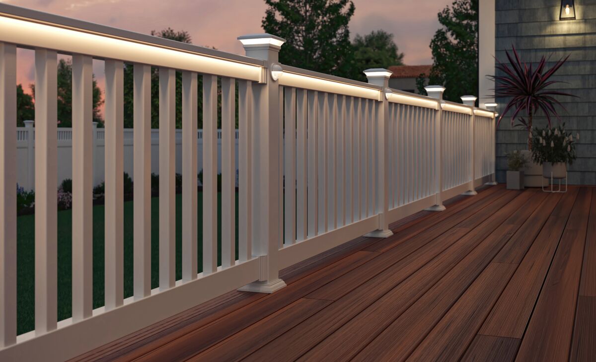 Una barandilla blanca bien iluminada bordea una terraza de madera al atardecer. La suave iluminación crea un ambiente acogedor. Una maceta y los árboles sugieren un entorno exterior tranquilo.