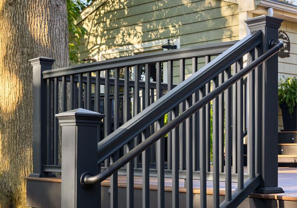 A black railing on a wooden deck is shown in sunlight, next to a tree trunk. It leads to a porch with plants. The scene is peaceful and inviting.